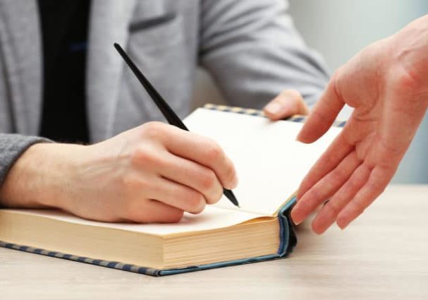 Author signing autograph in own book at wooden table on light blurred background